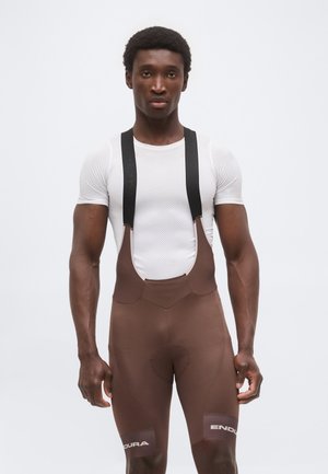 Mies pukeutuneena valkoiseen tekstuuri lyhythihaiseen paitaan ja ruskeisiin pyöräilysortsiköihin mustilla remmeillä, seisoo yksiväristä valkoista taustaa vasten.