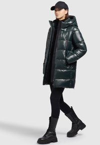 Donkergroen pufferjack met een glanzende afwerking, ritszakken, capuchon, en zwarte leggings gecombineerd met stevige zwarte veterschoenen.
