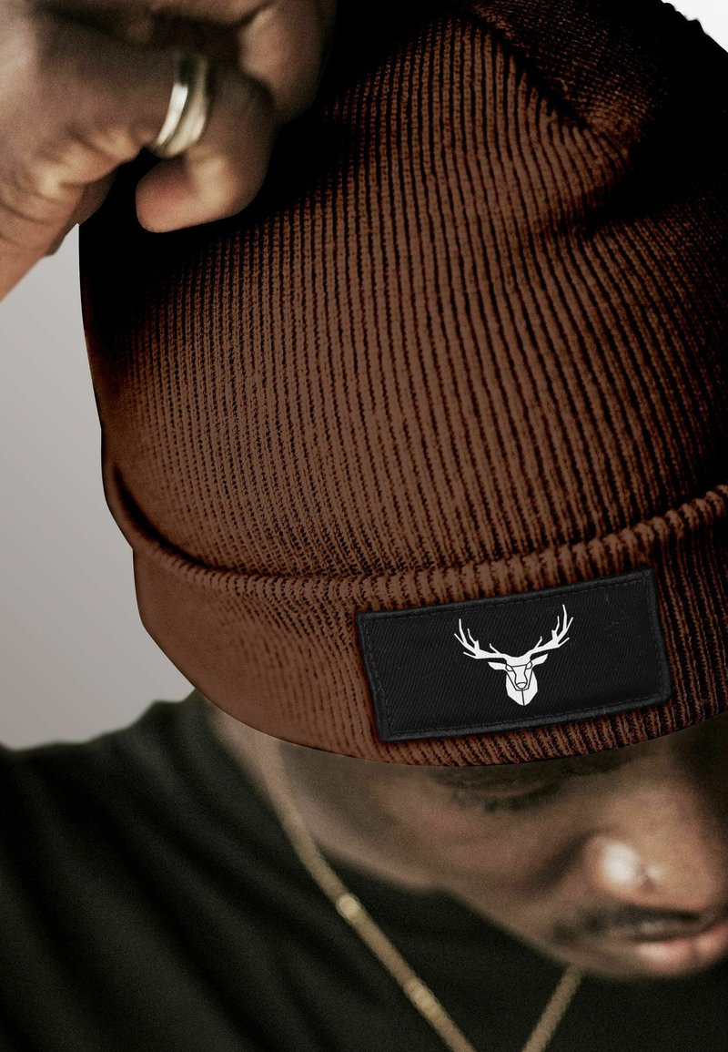 Brown ribbed beanie made of knit material, featuring a rectangular black patch with a white deer head logo on the front.