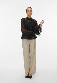 Černá průsvitná blůza s dlouhými rukávy a límcem na mašličku, kombinovaná s béžovými širokými kalhotami a černými špičatými lodičkami.