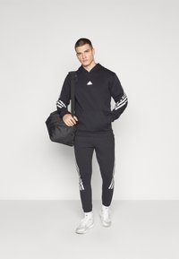 Felpa nera con logo Adidas bianco, strisce bianche a contrasto sulle maniche, pantaloni jogger neri abbinati e una borsa da palestra nera.