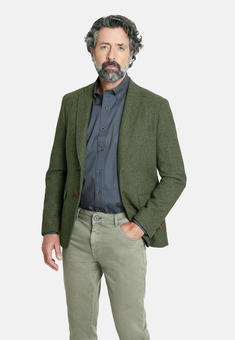 Blazer de lana verde con solapa muesca, tejido texturizado y dos botones; llevado sobre una camisa azul marino con un patrón de lunares y pantalones gris claro.