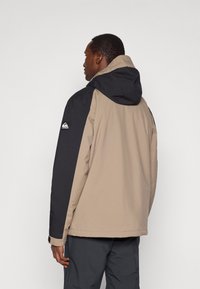 Άνδρας που στέκεται με την πλάτη στραμμένη, φορώντας μπεζ και μαύρο μπουφάν με κουκούλα και με λογότυπο στο αριστερό μανίκι, καθώς και σκούρες παντελόνες.