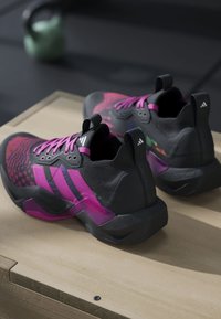 Ζεύγος μαύρα και ροζ αθλητικά παπούτσια Adidas σε ξύλινη επιφάνεια με θολωμένο kettlebell στο παρασκήνιο.