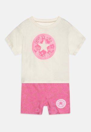 Camiseta de manga corta color crema con un logo de Converse en rosa, combinada con pantalones cortos rosas que presentan un estampado floral y un detalle de logo en el lado.