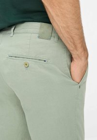 Pantaloni verde chiaro realizzati in un tessuto morbido, con una tasca sul retro con chiusura a bottone e un'etichetta con marchio sulla vita.