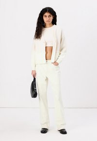Hvid, tekstureret cardigan over en hvid crop top, parret med lyse, løst siddende bukser og en sort, nittet håndtaske. Sorte sko.