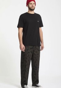 Zwart T-shirt met korte mouwen en een klein logo; donkergroene broek met een subtiel vleermuispatroon; zwarte sneakers; rode gebreide muts.