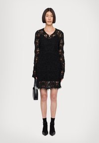 Abito in pizzo nero con maniche lunghe, intricate fantasie floreali, dettagli in perline, orlo scallop, abbinato a una borsa nera e scarpe a punta.