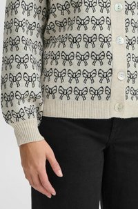 Cardigan en maille crème avec un motif de nœuds noirs répété, doté d'une fermeture à boutons et de poignets à côtes, porté sur un jean noir.