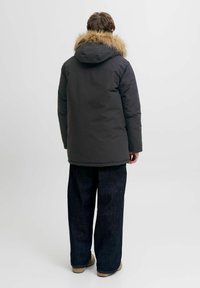 Sort dunjakke med en pelsforet hætte, tyk polstring og længere rygdesign, parret med løse, mørkeblå denimbukser og brune støvler.