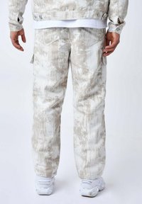 Pantalones cargo beige con un patrón abstracto texturizado. Presentan bolsillos laterales, corte recto y un ajuste relajado. Combinados con zapatillas blancas.