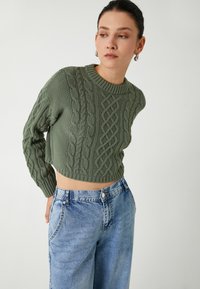 Grüner, gestrickter Cropped-Pullover mit Zopfstrickmustern, rundem Ausschnitt und langen Ärmeln, kombiniert mit hellblauen, hoch taillierten Jeans.