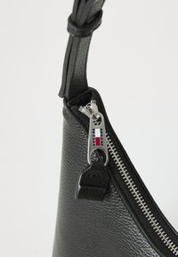 Bolso de mano de cuero negro con un acabado texturizado. Cuenta con una cremallera plateada y un tirador con la marca. Tiene forma curva y una correa ajustable.