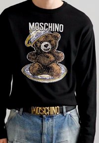 Suéter negro con un gráfico de un oso de peluche en un plato, con el texto "MOSCHINO" arriba. Cinturón plateado y dorado con el logo "MOSCHINO".