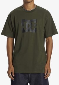 Olive zelené bavlnené tričko s čiernym logom DC a hviezdou. Krátke rukávy, okrúhly výstrih a voľný strih.