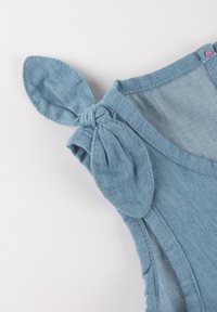 Hellblaues Denim-Oberteil mit einer verdrehte Knoten- und Ohren-Detail an den Schulterträgern, sichtbaren Nähten und weicher Textur.