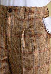 Rjave kariraste hlače z teksturiranim tkanjem, okrašenim z modrimi, rdečimi in črnimi črtami, z enim gumbom za zapiranje in stranskimi žepi.