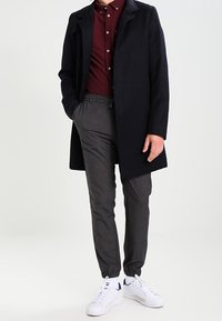 Cappotto nero, camicia a bottone bordeaux, pantaloni grigi a sigaretta e sneakers bianche. Le caratteristiche includono un colletto, tasche e polsini elastici.