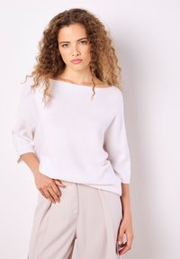 Weißer Strickpullover mit weitem Bootsausschnitt und drei Viertelärmeln, kombiniert mit beigen Hosen. Glatte Textur, lässige Passform.