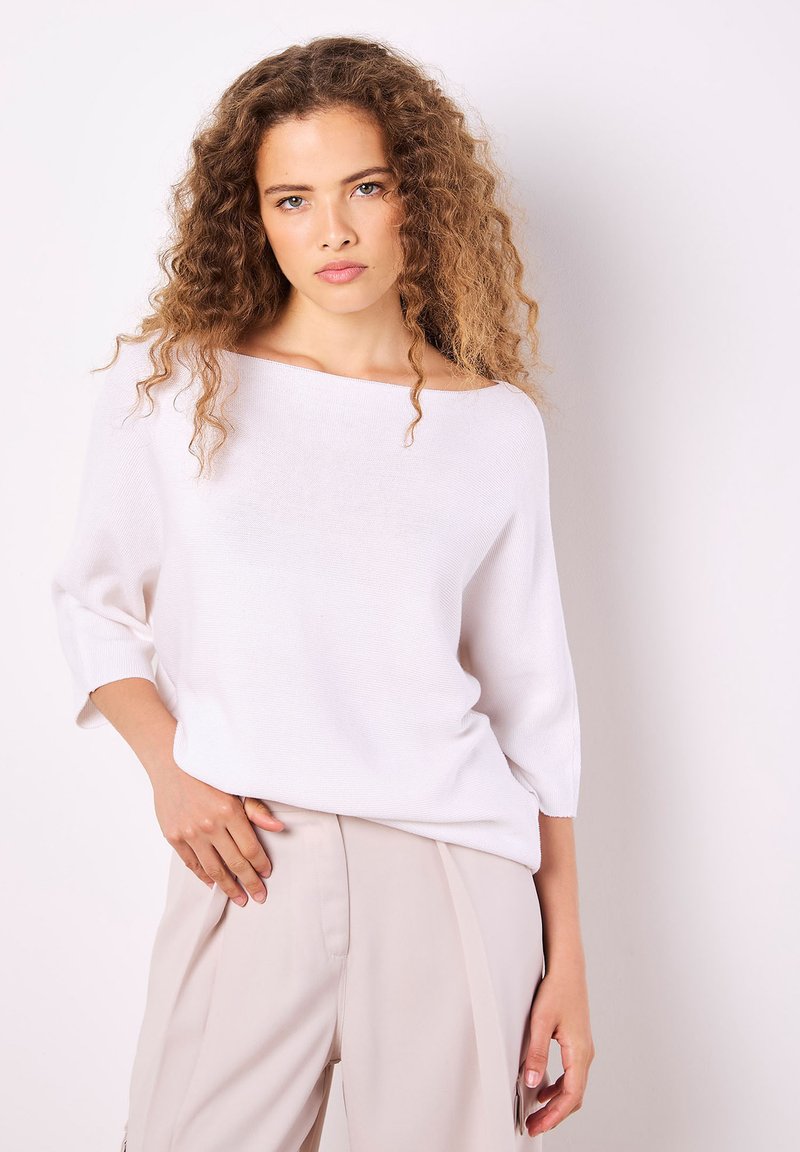 Weißer Strickpullover mit weitem Bootsausschnitt und drei Viertelärmeln, kombiniert mit beigen Hosen. Glatte Textur, lässige Passform.