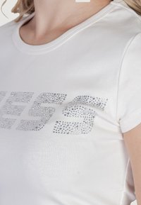 Balta medvilninė marškinėlių su trumpomis rankovėmis ir apvaliu iškirpimu, puošta dideliu blizgančių cirkonijų dizainu priekyje. Lygus paviršius.