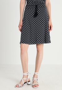 Robe à motifs noir et blanc avec une taille nouée, présentant un design géométrique. Associée à des sandales blanches à brides et à de petits talons carrés.