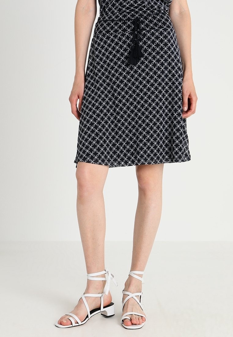Robe à motifs noir et blanc avec une taille nouée, présentant un design géométrique. Associée à des sandales blanches à brides et à de petits talons carrés.