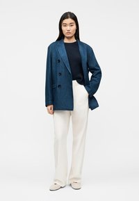 Γυναίκα που φοράει μπλε σκούρο σακάκι διπλής σειράς κουμπιών, μαύρο τοπ, λευκό παντελόνι με φαρδιά πόδια και μπεζ παπούτσια, στέκεται με το ένα χέρι στην τσέπη.