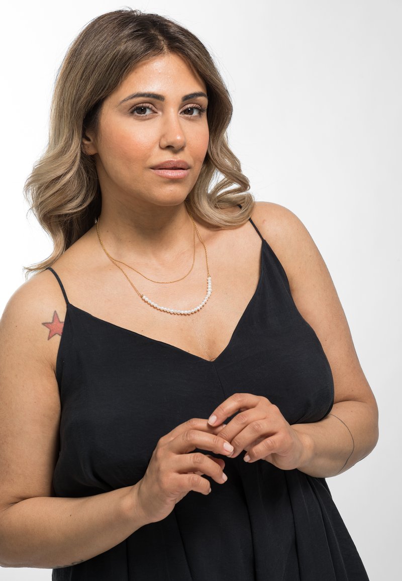 Collier en or superposé avec des perles blanches, porté sur une robe noire sans manches. Texture lisse et design simple avec un décolleté en V.