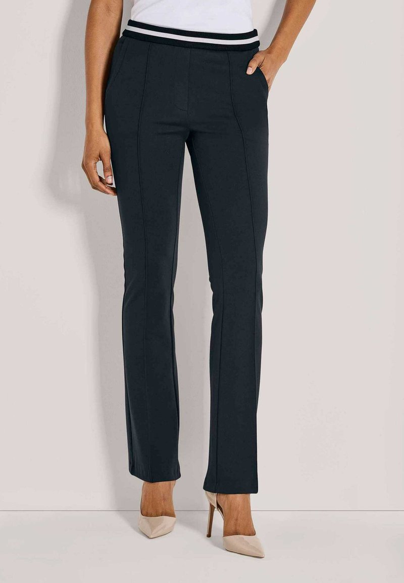 Vrouw in zwarte, op maat gemaakte, rechte broek met voornaden, handen in de zakken, gecombineerd met beige, puntige hakken.