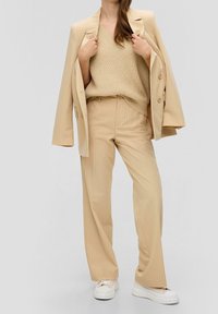 Beige gestreepte kostuum met een gestructureerde blazer, ontspannen fitbroek en een lichte gebreide v-hals trui. Gecombineerd met witte chunky sneakers.