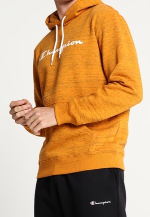 Person iført en orange Champion hættetrøje med hvide snore og sorte Champion sweatpants foran en ensfarvet baggrund.