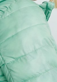 Chaqueta acolchada verde claro con textura acolchada, con costuras horizontales y cierre con cremallera en el cuello. Material suave y brillante.