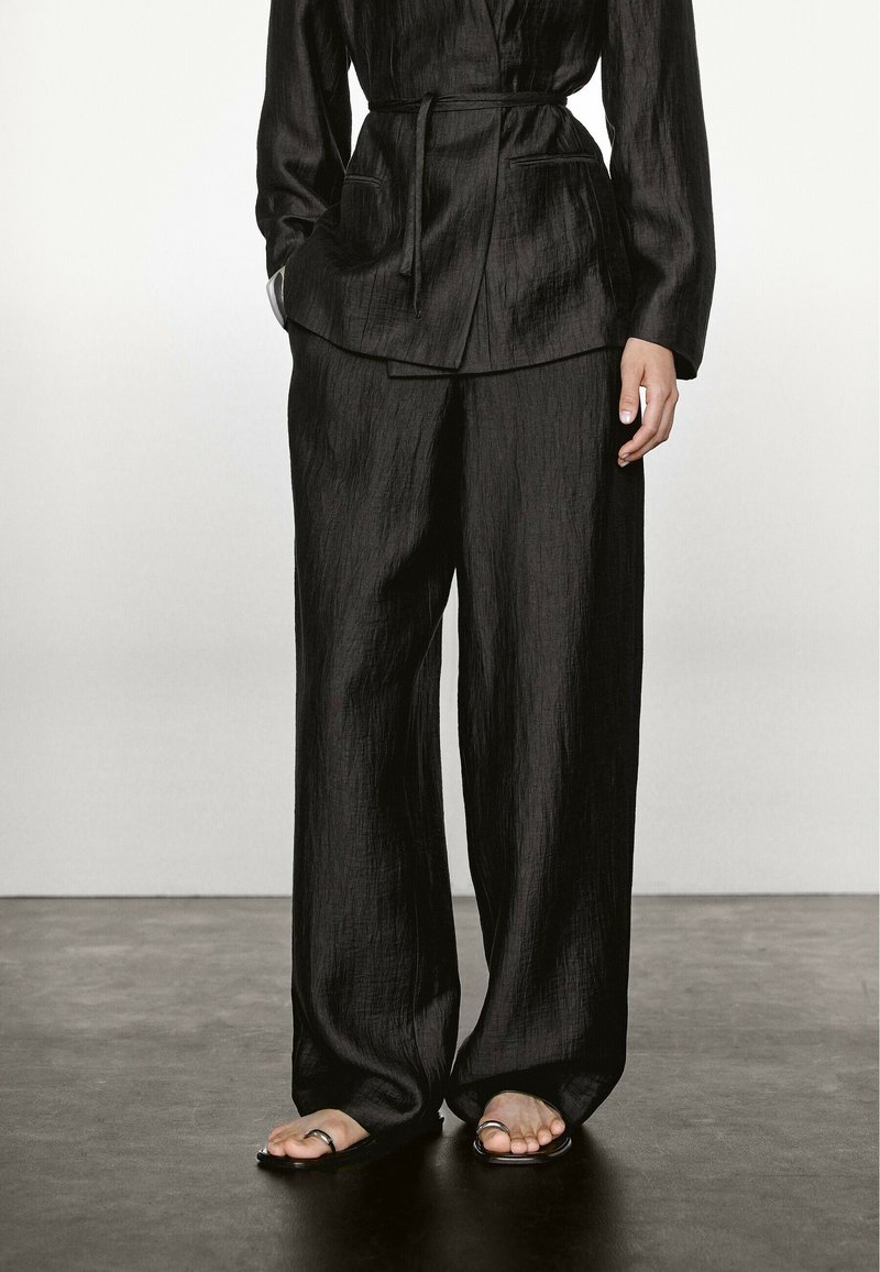 Una persona sta in piedi indossando un completo nero testurizzato con una giacca cintrata e pantaloni larghi, abbinati a semplici sandali su un pavimento scuro.