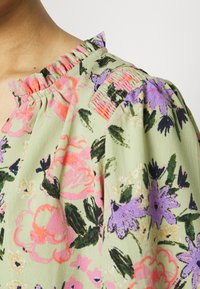 Gros plan sur un chemisier à col volanté avec un motif floral rose, violet et vert sur un tissu vert clair.