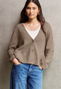 Brun stickad cardigan med V-ringning, fyra knappar och ribbade ärmslut, tillsammans med en vit topp och blå denimjeans. Texturerad tygdetalj synlig.