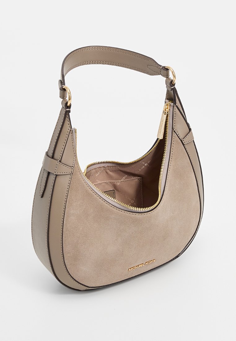 Beige suède schoudertas met een gebogen vorm, leren accenten, gouden rits en een binnenzak. Merkennaam is op de voorkant gedrukt.