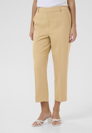 Beige maßgeschneiderte Hosen mit schmal zulaufendem Bein, die vordere Falten und eine glatte Oberfläche aufweisen, kombiniert mit weißen, schmalen Blockabsatzsandalen.