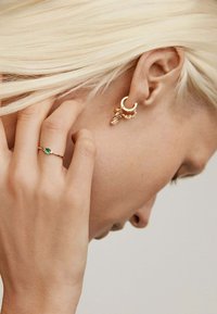 Pendientes de aro dorados con acentos colgantes texturizados y un anillo delicado que presenta una piedra preciosa verde, todo contra un fondo neutro.