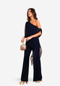 Combinaison navy à une épaule drapée, avec un superposé fluide et un design à jambes larges. Associée à une pochette métallique et des talons compensés.