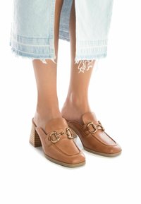 Mules de cuero tan con tacón elevado, que presentan un detalle de hebilla en tono dorado y acentos bordados. Llevados con una falda de mezclilla clara.