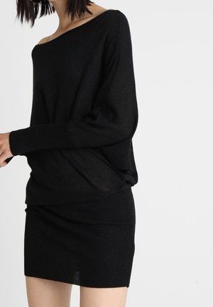 Woman wearing an off-shoulder black long-sleeve sweater and a matching black ribbed mini skirt against a plain white background.