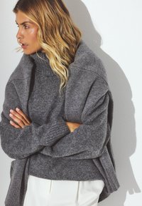 Suéter de gran tamaño en color gris con cuello alto, confeccionado en suave tejido de punto, que presenta detalles acanalados en los puños y el dobladillo, combinado con pantalones blancos.