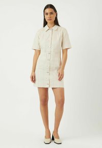 Robe à manches courtes, de couleur claire, présentant un design à boutons sur le devant, un col pointu et des détails de couture contrastants. Portée avec des chaussures à talons carrés.