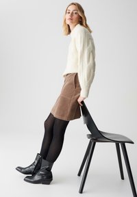 Maglione chunky in crema lavorato a maglia, gonna in velluto a coste marrone, collant neri e stivaletti neri. La modella posa accanto a una sedia nera in un ambiente minimalista.