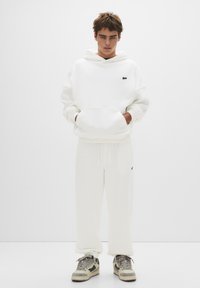 White hoodie with a front pocket and a small black logo, paired with white sweatpants. Model wears grey and black sneakers. Minimalist style.
