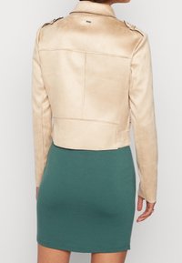 Veste courte en cuir synthétique beige avec épaulettes, coutures verticales et texture lisse, portée sur une robe verte ajustée.