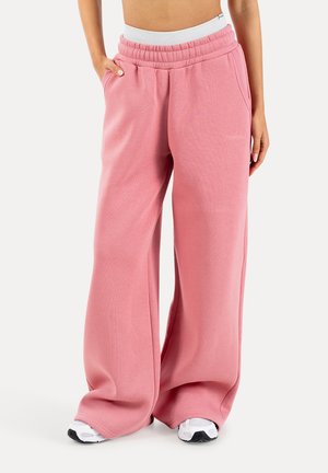 Persoon die losse roze joggingbroek met elastische tailleband en witte sneakers draagt, staand met één hand in de zak tegen een witte achtergrond.