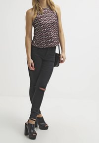 Zwarte hoge taille skinny jeans met versleten knieën, gecombineerd met een mouwloos, gestreept topje in een wijnrode en crèmekleurige zigzagpatroon. Chunky zwarte hakken.